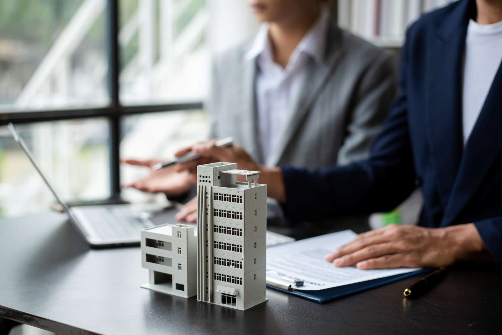 Menschen in Anzügen sitzen an einem Tisch, Miniaturgebäude steht vor ihnen
